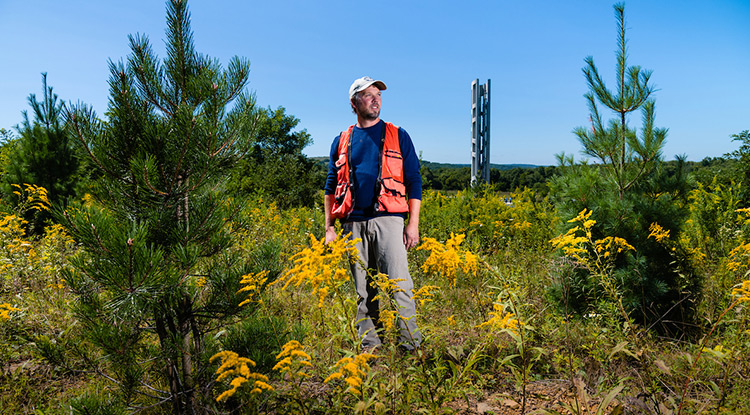 Michael Tyree stands among young pine trees surrounded by goldenrod in a field around the Flight 93 National Memorial. A tall structure known as the Tower of Voices is visible behind him.