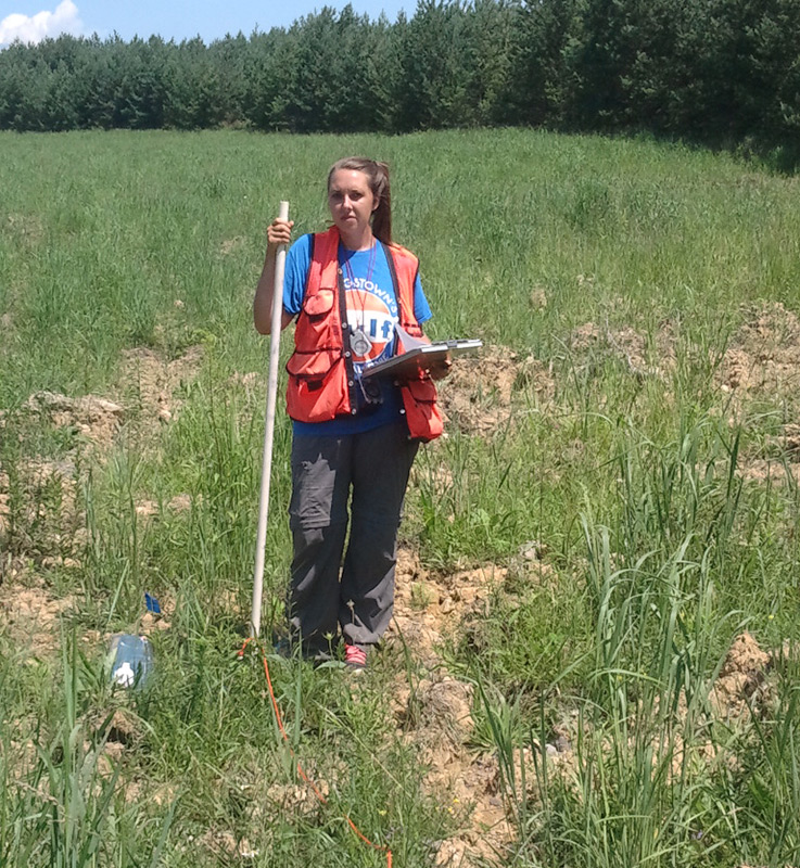 Shannon Johns Pierce holds a piece of white PVC pipe in a field with pine trees in the background as she works at the Flight 93 memorial site. Part of a formation of PVC pipe is visible in the foreground.