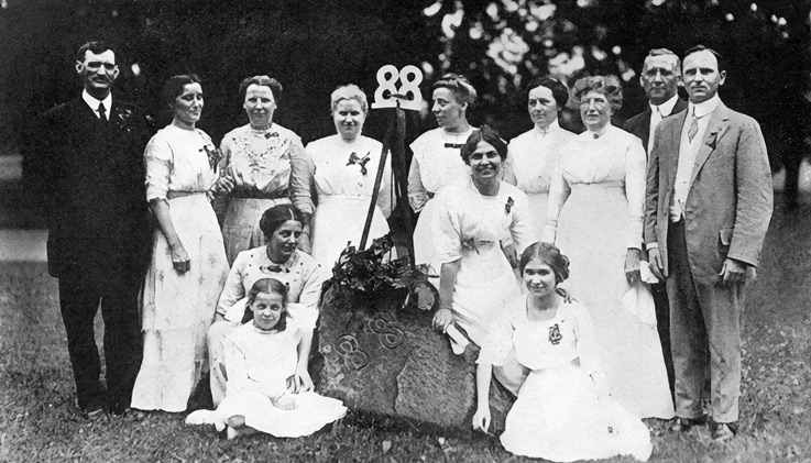 A group of men and women pose for an outdoor photo as they surround a rock engraved with “’88”