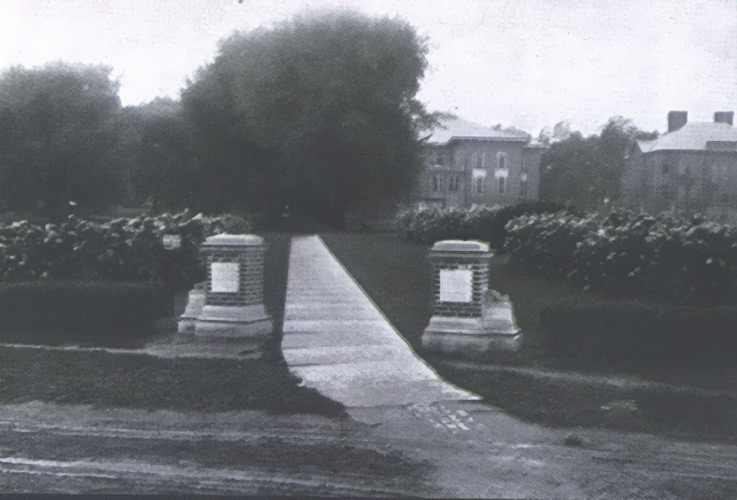 An old photograph of two brick markers on either side of a walkway that leads into the campus