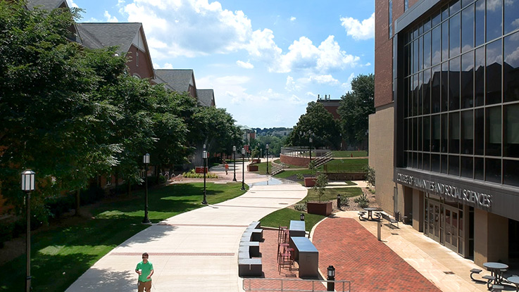 A student travels a broad walkway that snakes through campus green space, with buildings on either side