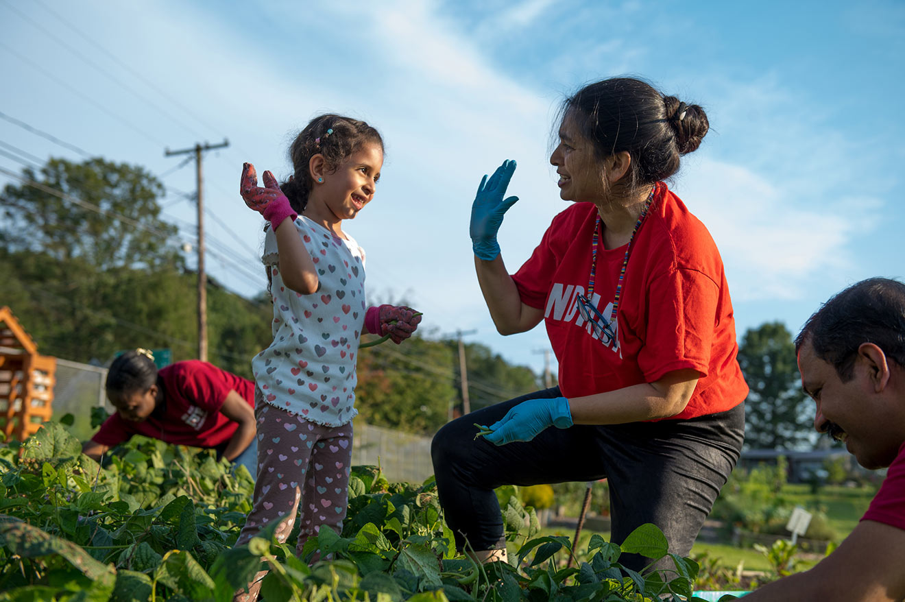 A young girl in gardening gloves and a woman resting on one knee hold up their hands to do a high five. Two other people working in the garden can be seen next to them.