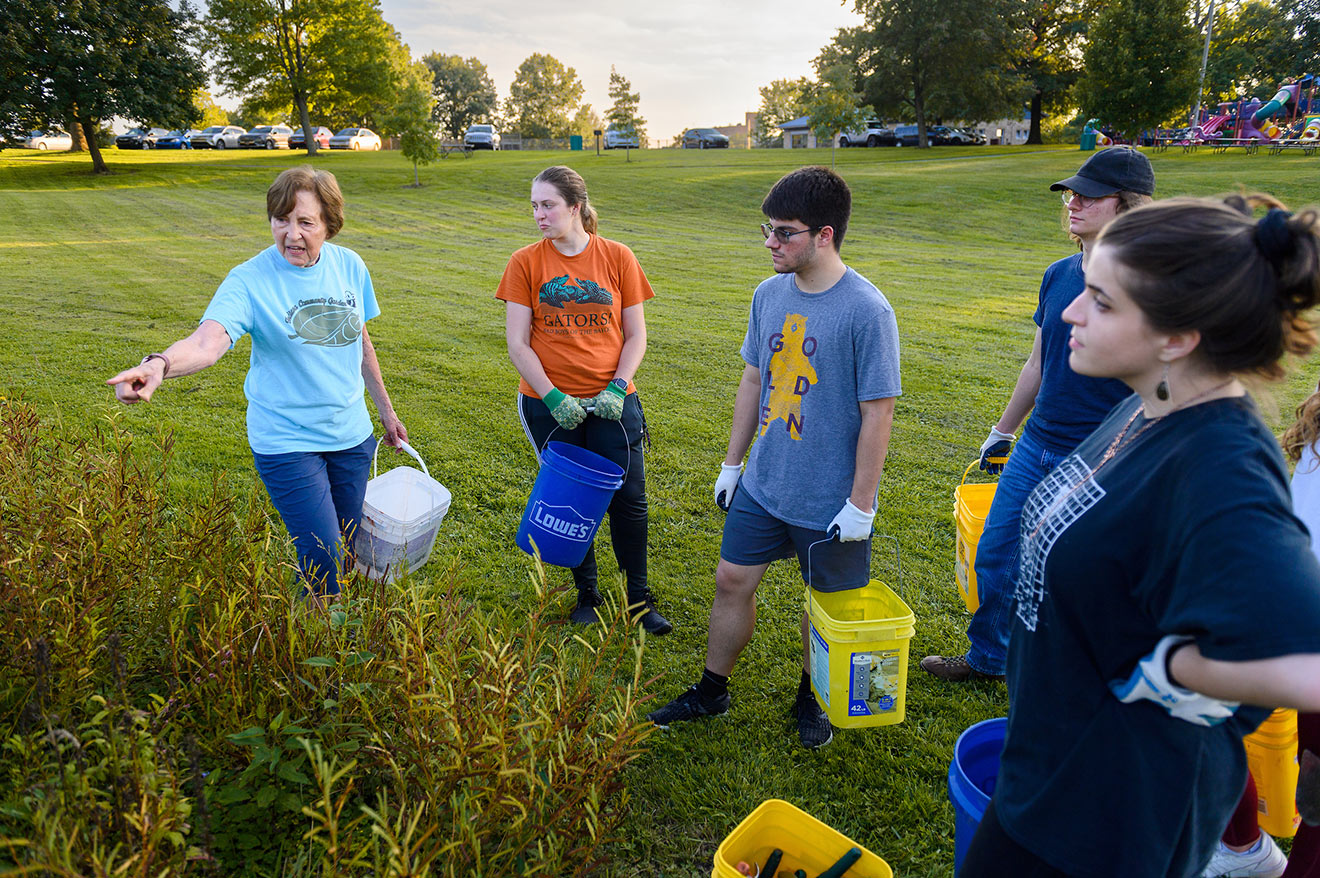 A woman holding a white bucket points to a garden with tall wildflowers, giving instructions to two young women and two young men who are holding blue or yellow buckets.