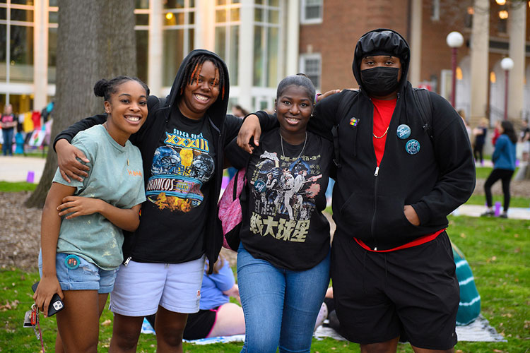 Four students smile and pose in the Oak Grove, standing side by side with their arms around one another’s shoulders. People sitting on a blanket, a tree trunk, several people milling about, and the front of the IUP Performing Arts Center are visible in the background.