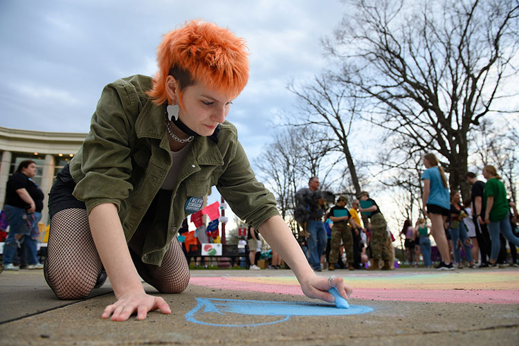 A ground-level view of a student with spiky reddish hair, who is on her knees, drawing a heart in light-blue chalk on an Oak Grove sidewalk. Several people and the IUP Performing Arts Center are visible behind her.