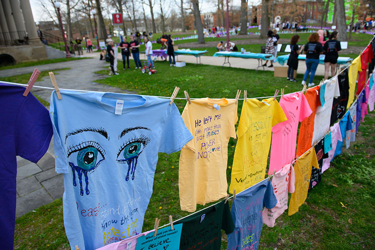 Colorful T-shirts with messages and designs hang on two clotheslines running close to parallel in the Oak Grove, with a corner of Waller Hall’s porch, display tables, and people standing and talking to one another in the background. A light-blue T-shirt decorated with tears streaming from two large eyes is prominent in the foreground.