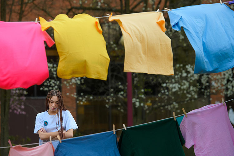A student looks at an assortment of colorful T-shirts hanging on two clotheslines in front of her, with trees and a building in the background.