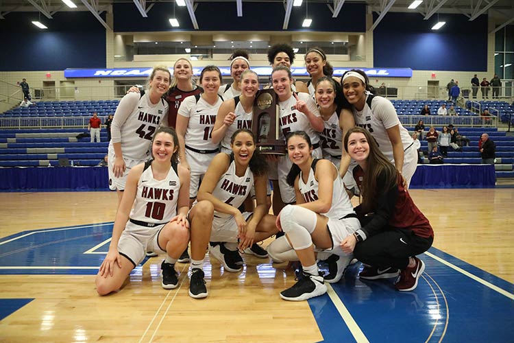 Fourteen young women, four kneeling in front, and most wearing white jerseys with “HAWKS” across the front, smile as they hold up an NCAA trophy while posing in the middle of a basketball court in an arena.