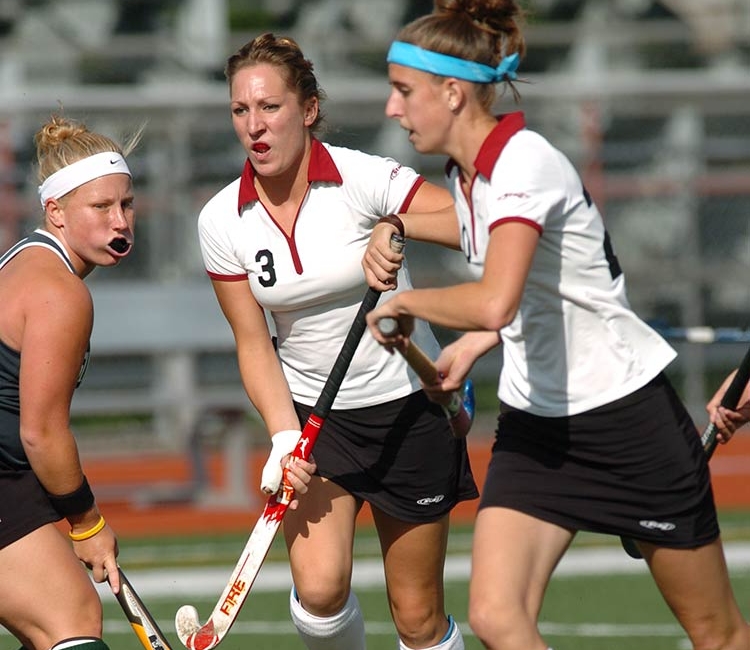 Two field hockey players in white jerseys with red collars and black skirts hold sticks as they run down the field while a player in a dark green jersey is turned toward them.