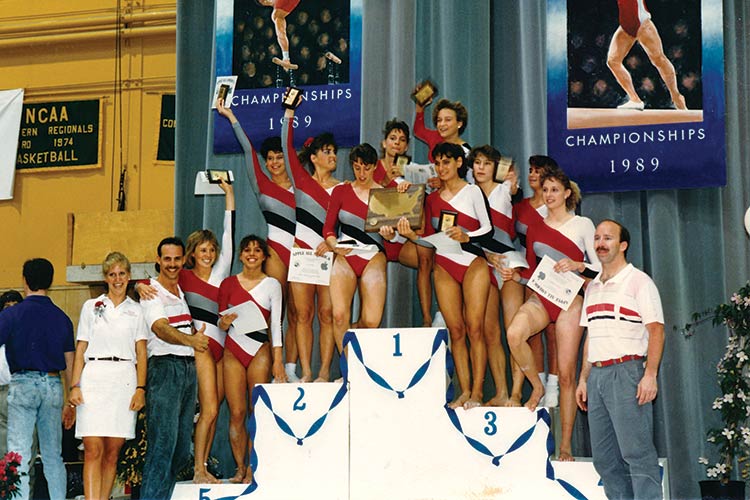 Eleven young women in leotards with wide red, gray, blue, and white diagonal stripes hold up small plaques and certificates as they crowd a podium for medal winners, with two men and another woman standing nearby.