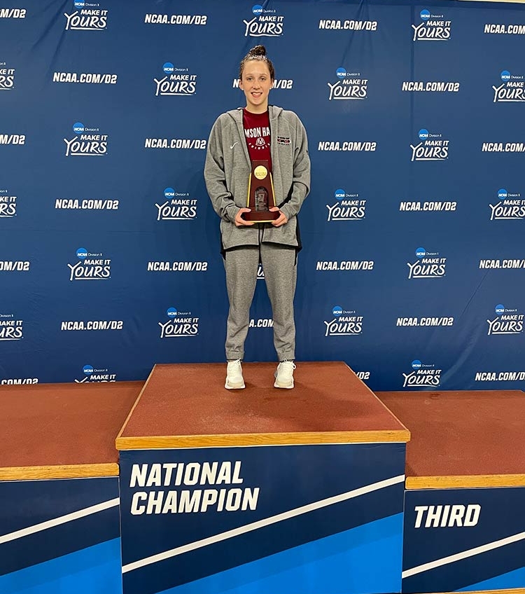 A young woman in a red Crimson Hawks T-shirt and gray warm-ups, with her hair in a high bun, smiles and holds an NCAA trophy as she stands at the top of a podium, labeled “National Champion.”