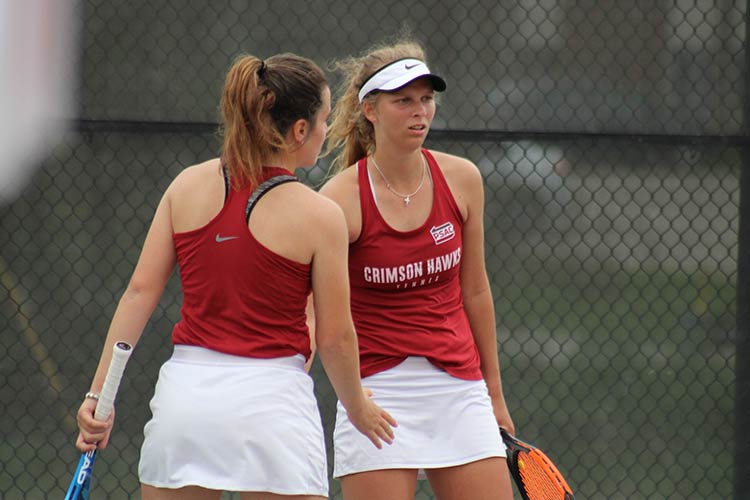 Two young women wearing red Crimson Hawks tank tops and white tennis skirts and holding rackets prepare to give a low hand slap while on the court.