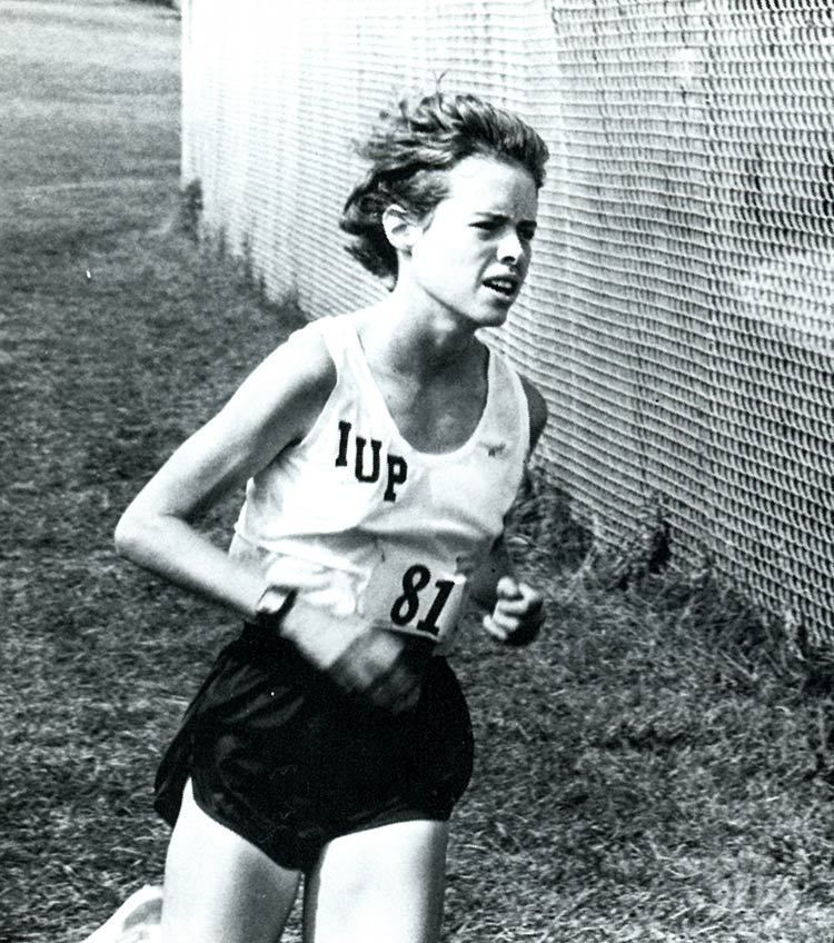 A young woman wearing a white IUP tank top and black running shorts runs in the grass outside a track fence.