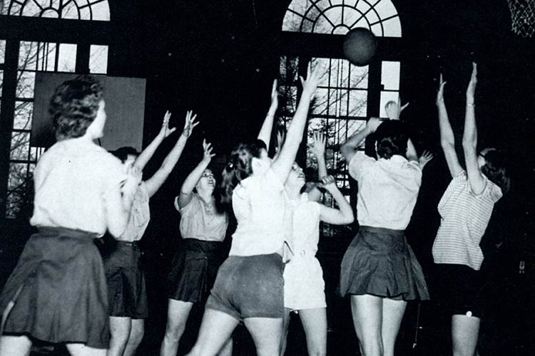 In this black and white photo, seven young women in shorts or athletic skirts reach up for a ball in a dark gymnasium, with sunlight and trees visible through arched windows in the background.