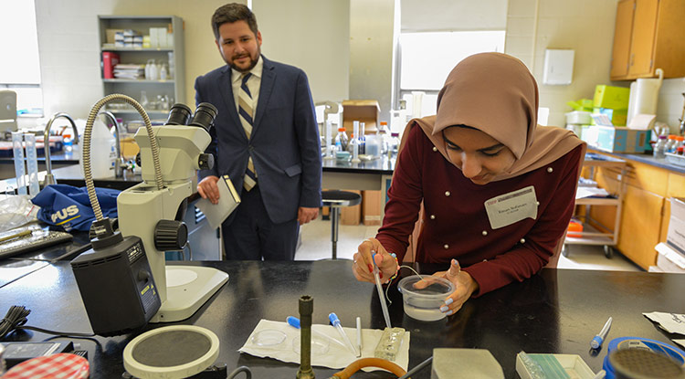 a young woman leaning over a lab table working on research while a man in a suit observes