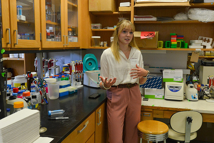a young woman talking and standing in the corner of a lab facing off camera