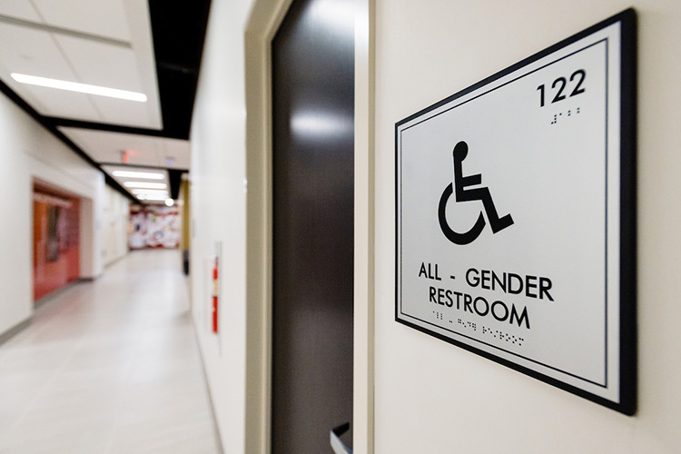 Hallway view of a sign with a wheelchair symbol and the words “ALL-GENDER RESTROOM” beneath it on a wall next to a black door