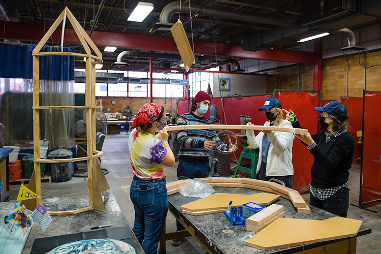 In a vast art studio setting and standing next to a wooden rocket frame, three people with face coverings hold up a semicircular wood cutout while a fourth person holds a small clear dome beneath it