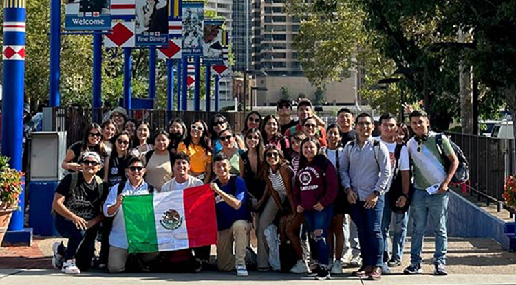 A large group of students smiling for the camera on a street while holding up a Mexican flag