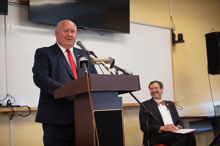 Congressman Glenn Thompson stands and speaks into a microphone at a podium in the front of a classroom setting with a whiteboard and a monitor behind him. IUP President Michael Driscoll, seated behind and to the side of the congressman, smiles as he looks to his right.  