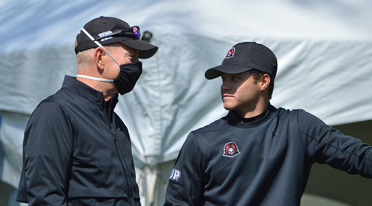 Dan Braun talks with IUP golfer Colin Walsh in front of a white tent on the sidelines of a golf tournament