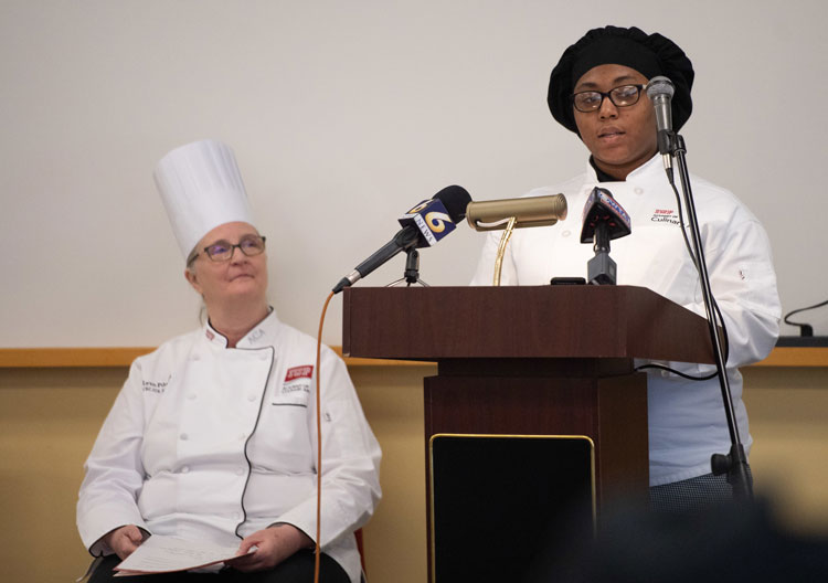 Shakyra McNeal, wearing a black chef’s cap and a white chef’s coat, stands and speaks at a podium with three microphones, some from news outlets. Chef Lynn Pike, wearing a white chef’s coat and hat, sits and listens behind and to the side of Shakyra. 
