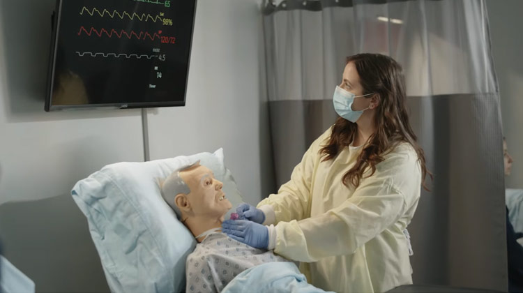 A nursing student working in a mock hospital room next to a bed containing a dummy dressed as patient