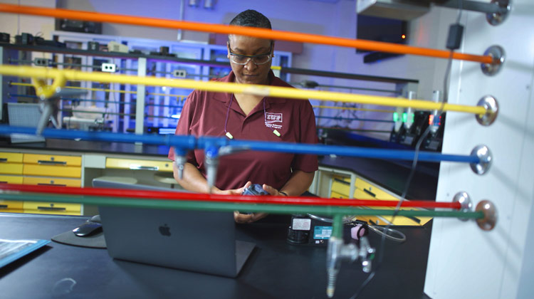 Cynthia Pugh in a lab working on a laptop behind a counter and multicolored pipes