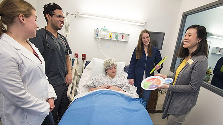 Smiling nursing students in a mock hospital room surround a bed containing a student dressed as a patient.  To the side an instructor is showing them multicolored learning materials.