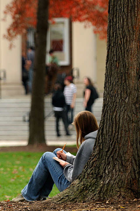 Student sitting with her back against a tree in the Oak Grove. The steps of Waller Hall can be seen in the background.