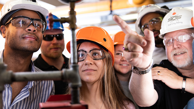 Students in hard hats look at Safety Science equipment as a professor explains it.