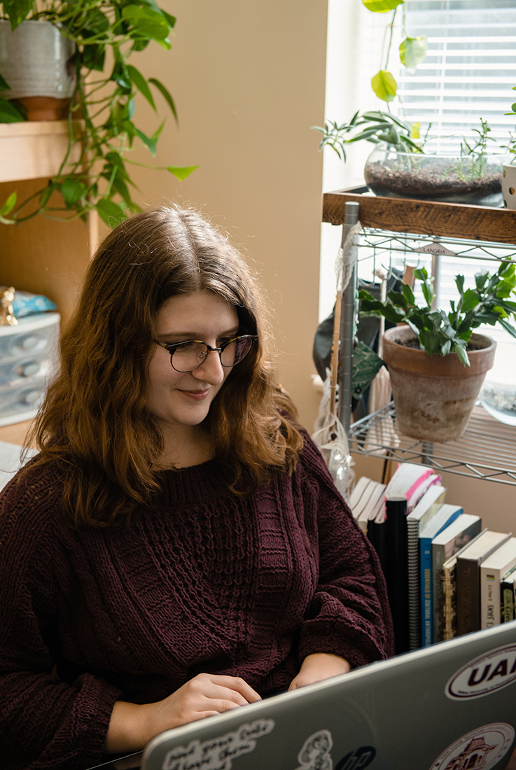 A student studies on her laptop in her room near a sunny window and some plants.