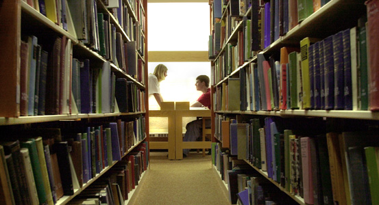 two students working at a table seen through a row of books at the library.