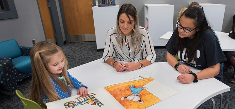Two students aspiring to be teachers work with a young child, who is looking at a picture book.