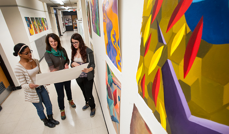 Two students talk with a professor about an art project in a hallway lined with colorful paintings.