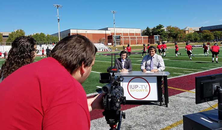 Students enrolled in COMM 360 – Digital Sports Production write, produce, and set up a pre-game production on the field during warm-ups of an IUP football game. Students enrolled in COMM 360 – Digital Sports Production write, produce, and set up a pre-game production on the field during warm-ups of an IUP football game.
