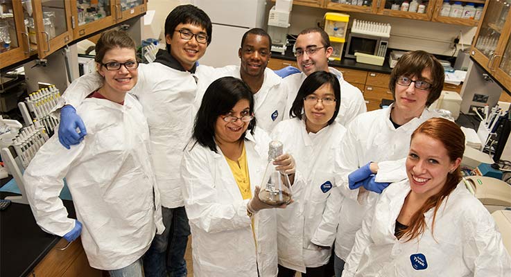 A group of students in lab coats pose for a photo with their professor in a lab
