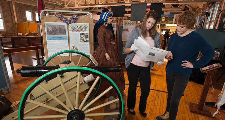 Two students look at documents while standing in a local museum near a small cannon Two students look at documents while standing in a local museum near a small cannon