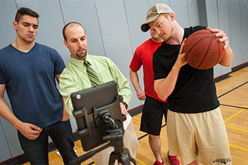 A Kinesiology professor instructs students from a laptop while one holds a basketball A Kinesiology professor instructs students from a laptop while one holds a basketball