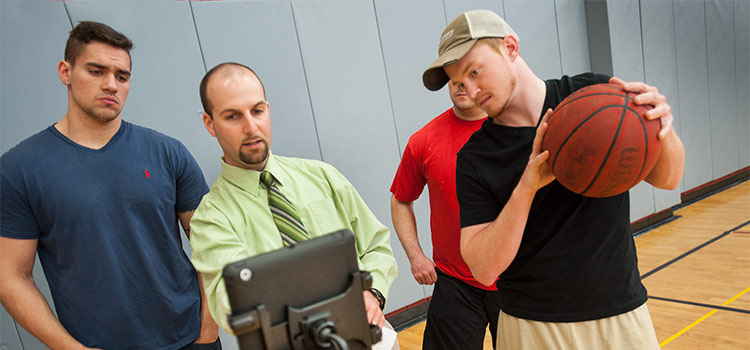Students in Kinesiology listen to a professor using a laptop as one student holds a basketball