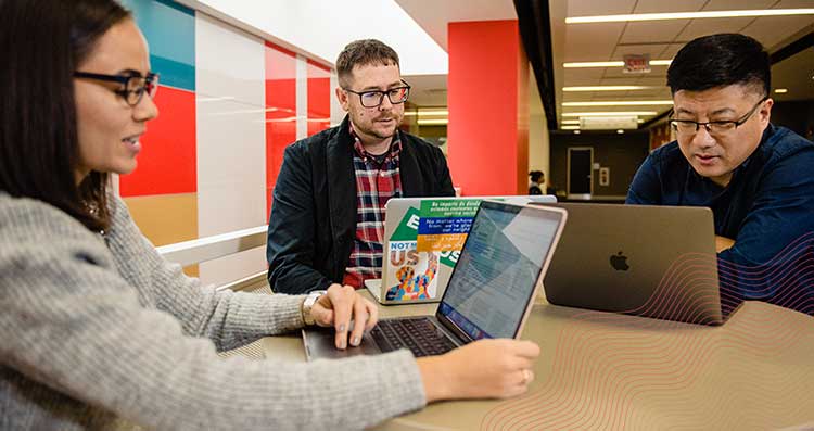 Three graduate students with laptops sit together at a table. 