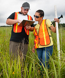Two students use surveying equipment in a field of tall grass to gather data.