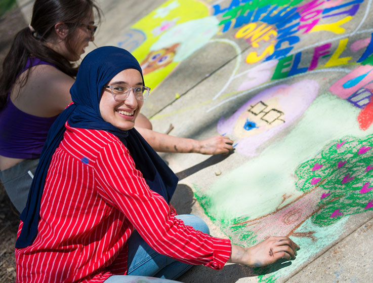 Two students working on their sidewalk chalk artwork, one of them smiling at the camera 