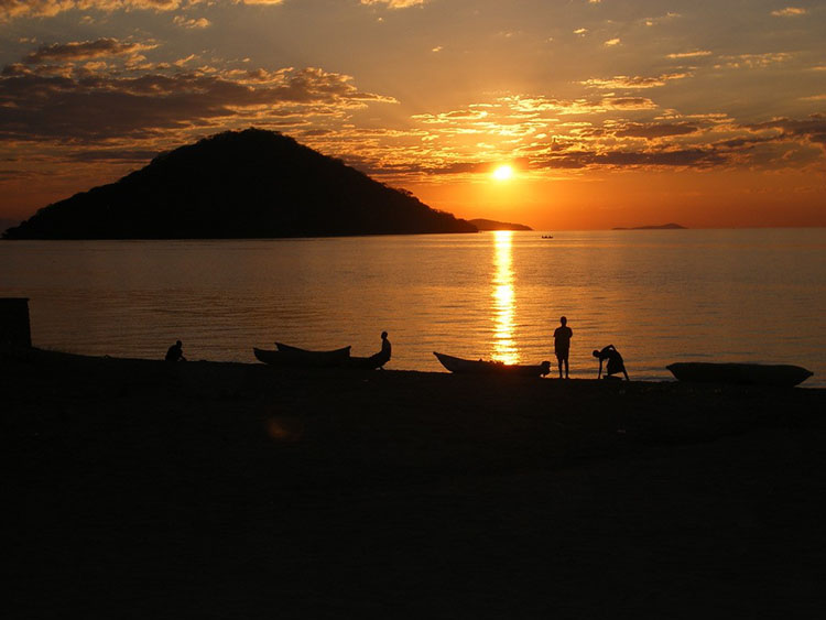 The sun rises or sets over water with people and boats in the foreground